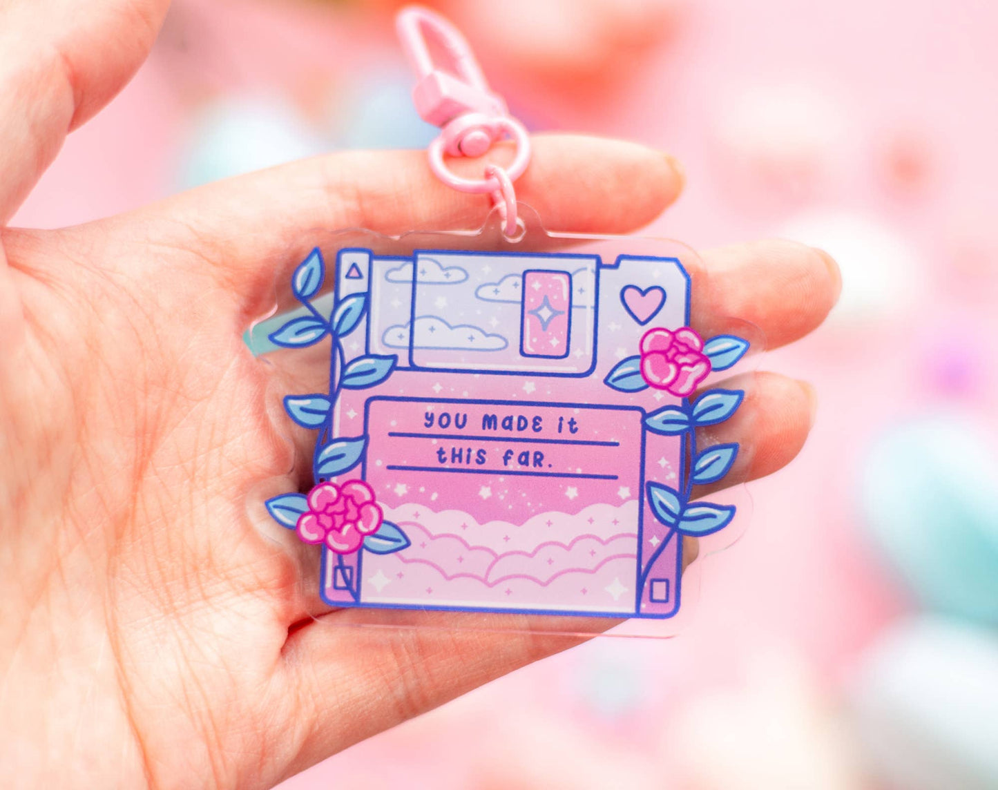A hand holding a translucent acrylic keychain charm shaped like a retro floppy disk, decorated with pink flowers, blue leaves, and the text 'YOU MADE IT THIS FAR.' on a soft pink blurred background.