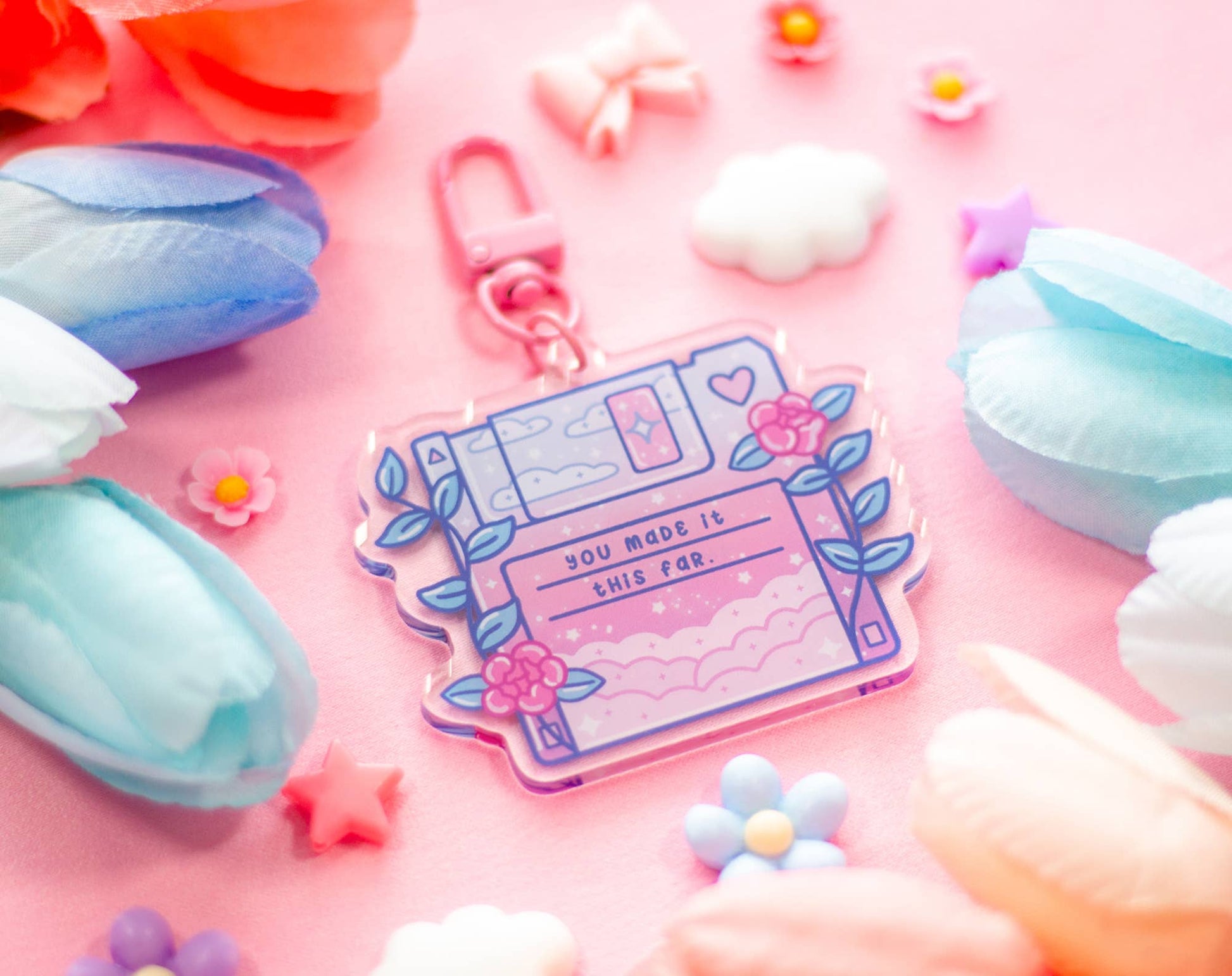 A pastel pink and lavender acrylic keychain charm shaped like a retro computer monitor, decorated with flowers, leaves, and the text 'YOU MADE IT THIS FAR,' photographed on a pink background with decorative paper flowers and clouds.