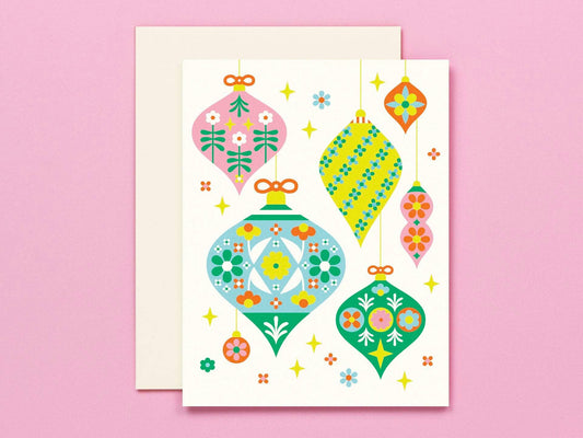 A square greeting card with a cream background featuring colorful retro Christmas ornament illustrations, shown with a matching cream envelope on a solid pink background.