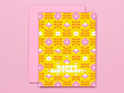 A retro-style 'Happy Birthday' greeting card with a yellow background, pink and white floral pattern, and a pink envelope behind it, set against a solid pink background.
