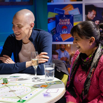 Two people laughing together at a table with a board game, surrounded by colorful posters.