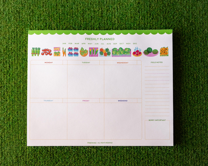 Weekly meal planner pad with colorful food icons on a green grass background