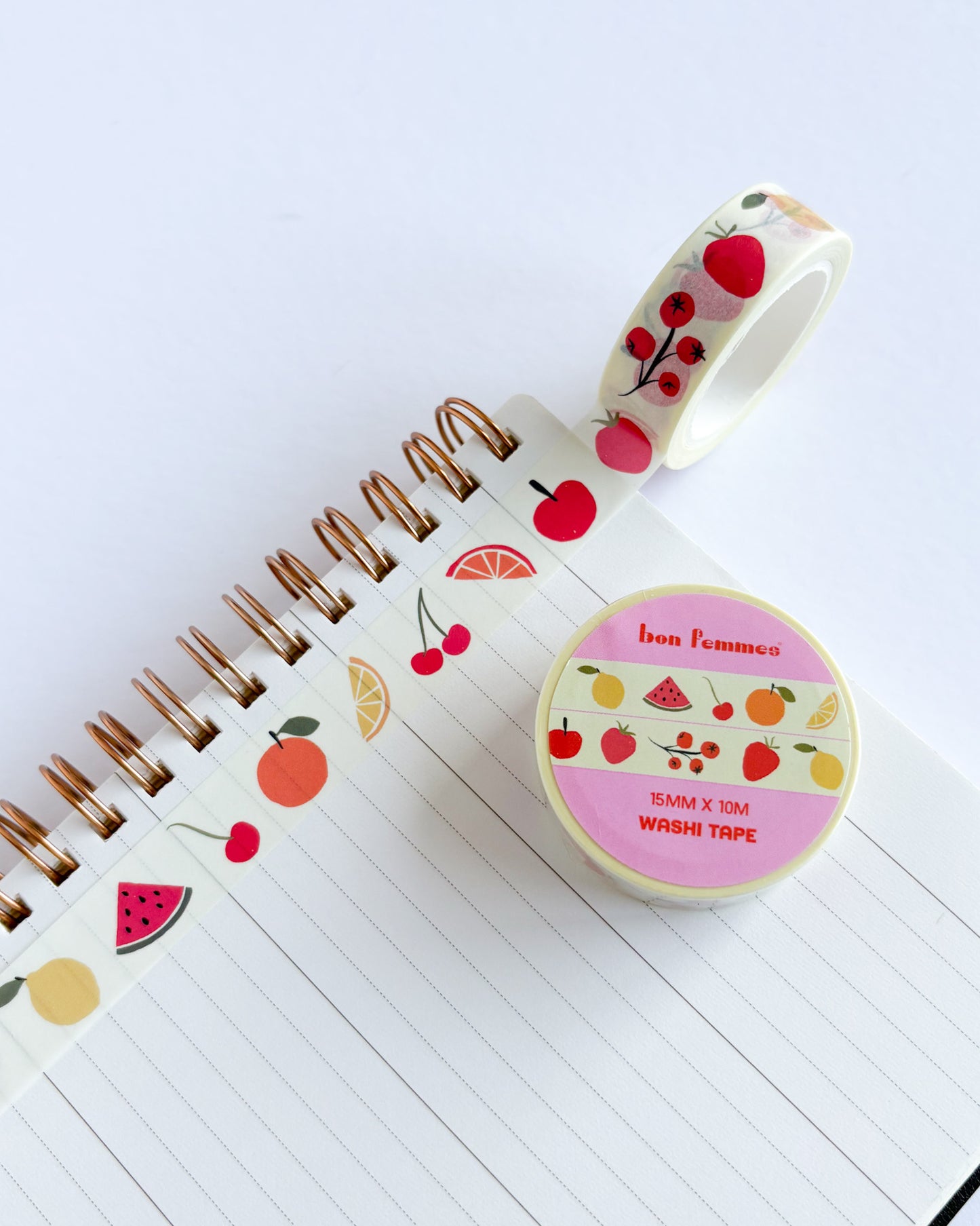 A roll of fruit-patterned washi tape and a spool of the same tape placed on an open lined notebook. The tape features illustrations of red cherries, strawberries, oranges, watermelon slices, and citrus on a white background.