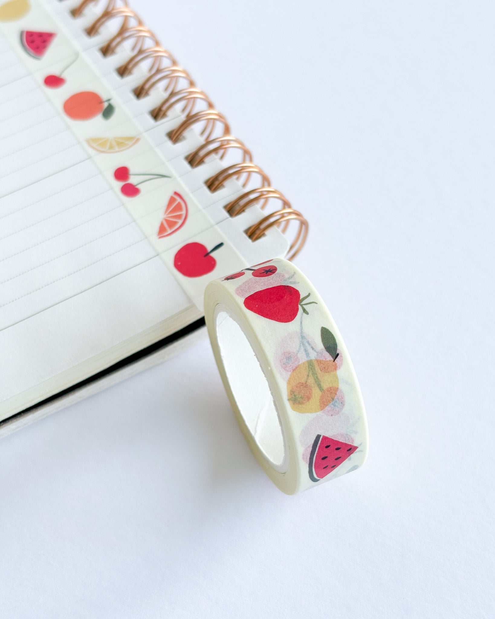 A roll of washi tape with a colorful fruit pattern, shown draped over an open spiral notebook on a clean white surface.