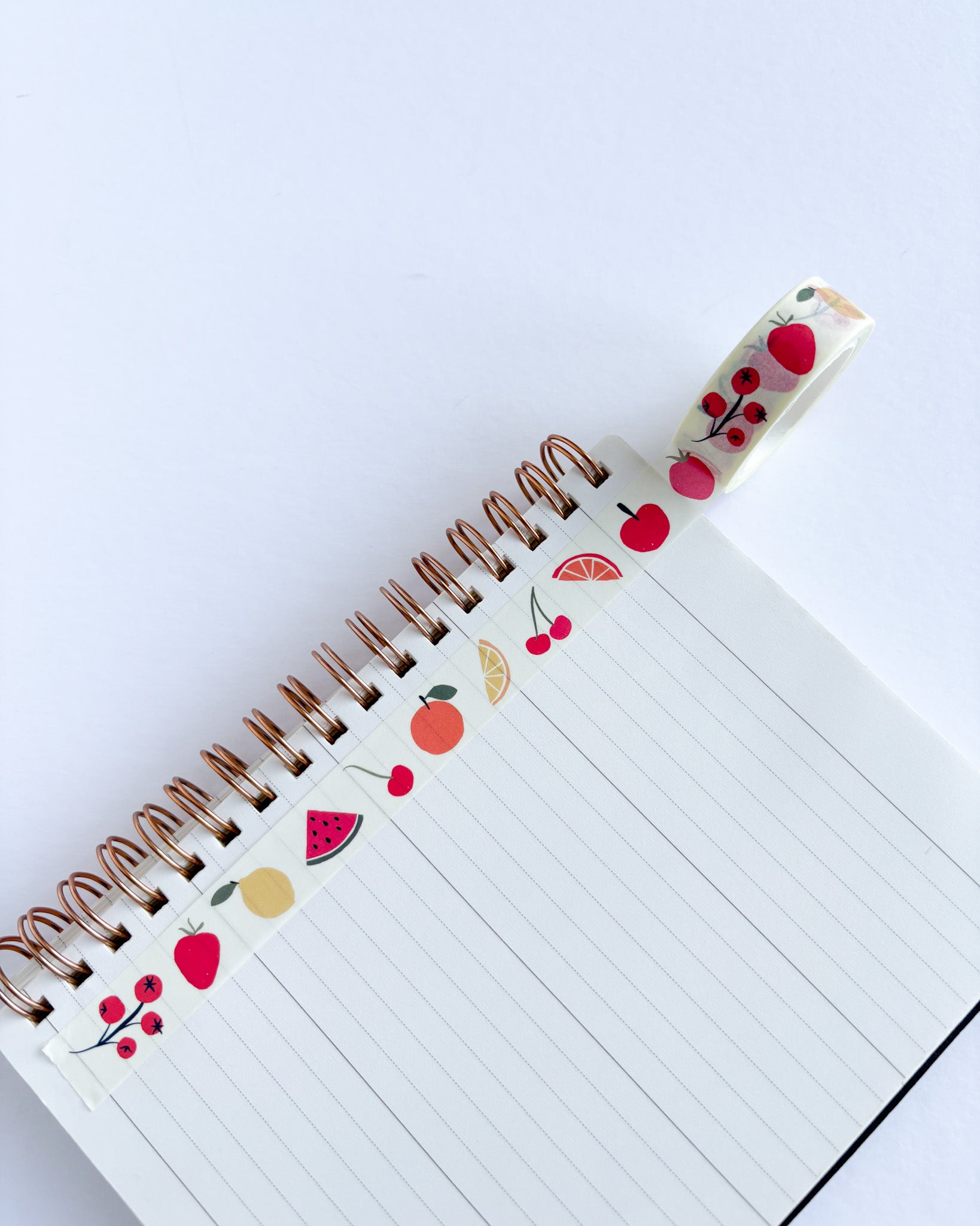 A spiral-bound lined notebook with decorative fruit-themed washi tape running along the binding. The tape features illustrations of red cherries, watermelons, oranges, and yellow citrus slices on a white background.