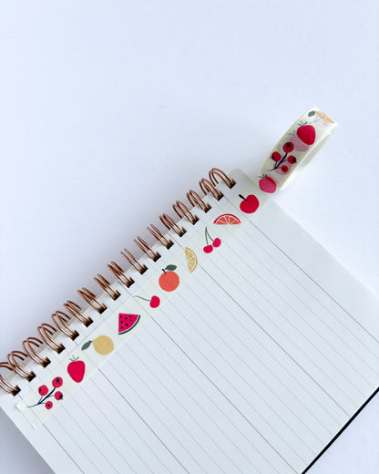 A spiral-bound lined notebook with decorative fruit-themed washi tape running along the binding. The tape features illustrations of red cherries, watermelons, oranges, and yellow citrus slices on a white background.