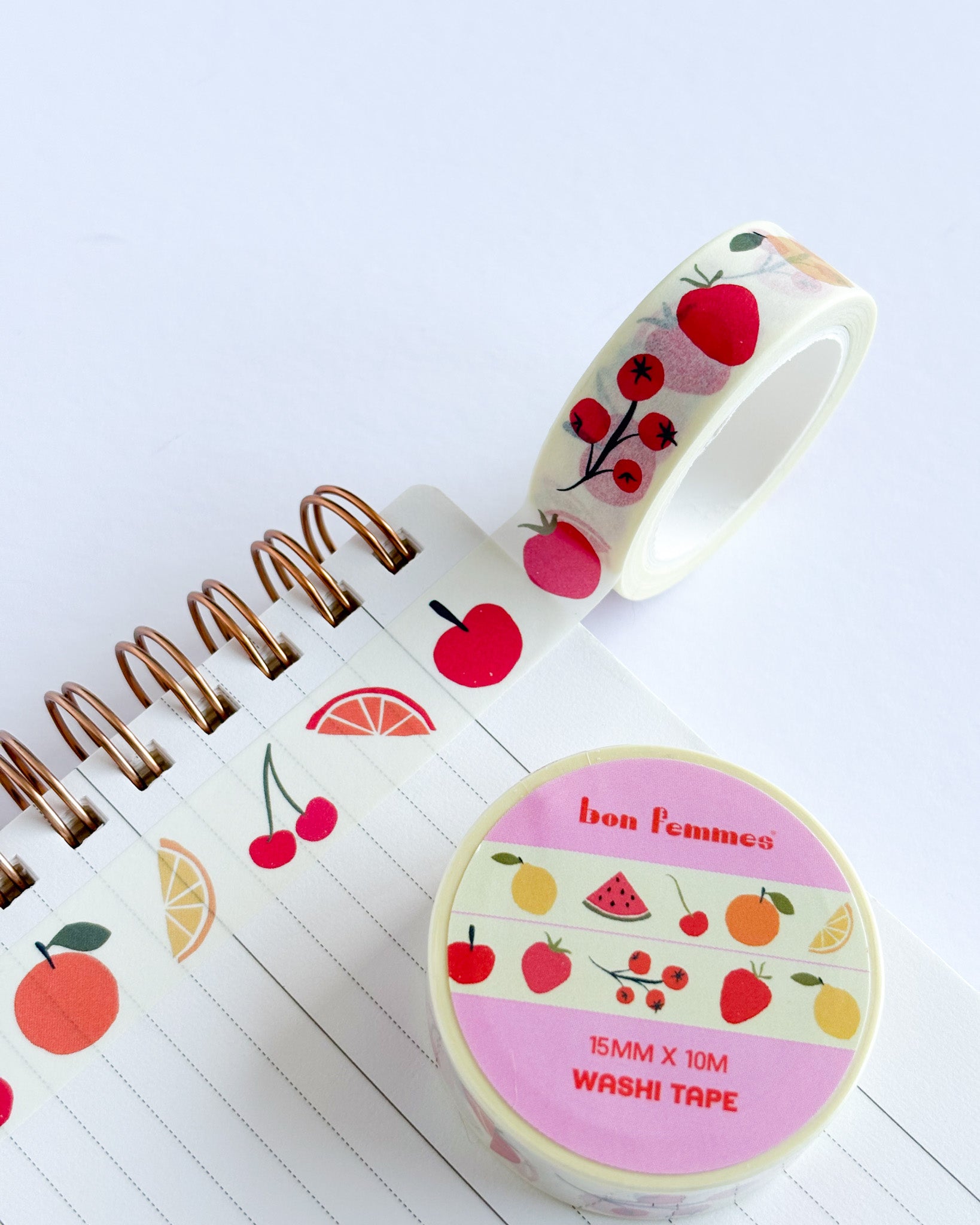 A roll of fruit-patterned washi tape and a matching spool labeled 'bon pommes' are shown on a white background, with some tape unrolled over lined paper next to a spiral notebook.