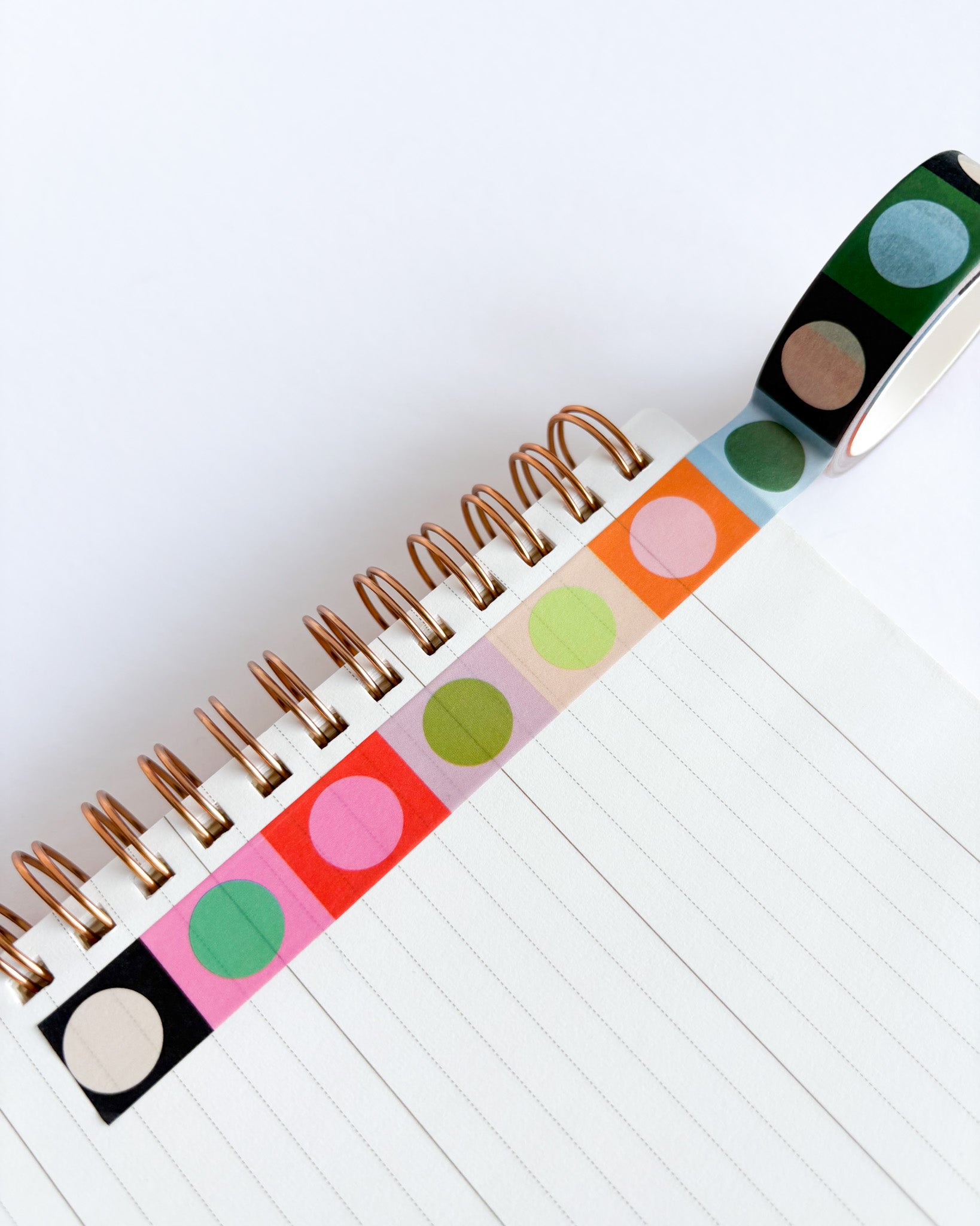 A roll of washi tape with colorful circular dots in black, green, blue, pink, orange, and light green, shown unrolled over a white spiral notebook.