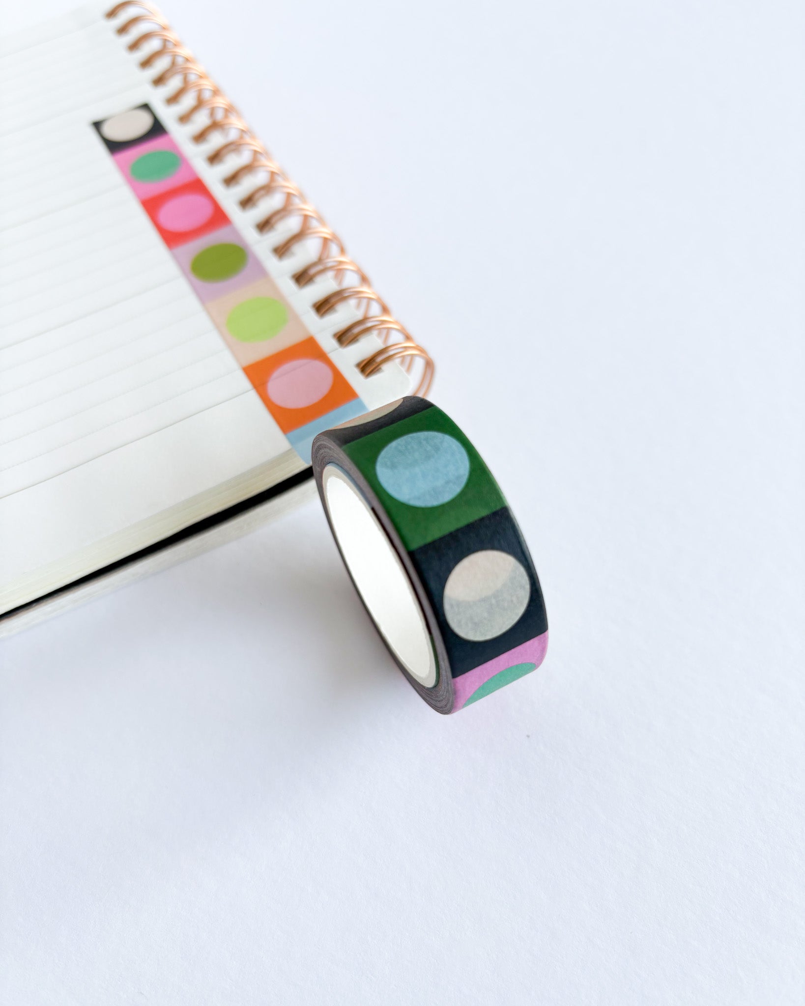 A roll of decorative washi tape with a black base and colorful circular dots in green, blue, pink, orange, and white, shown next to an open spiral notebook on a white surface.