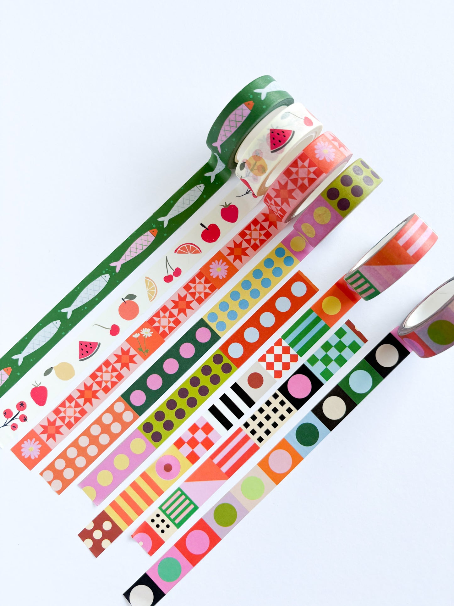 A set of colorful washi tapes with various patterns including fruit, fish, geometric shapes, stripes, and polka dots, arranged diagonally on a clean white background.