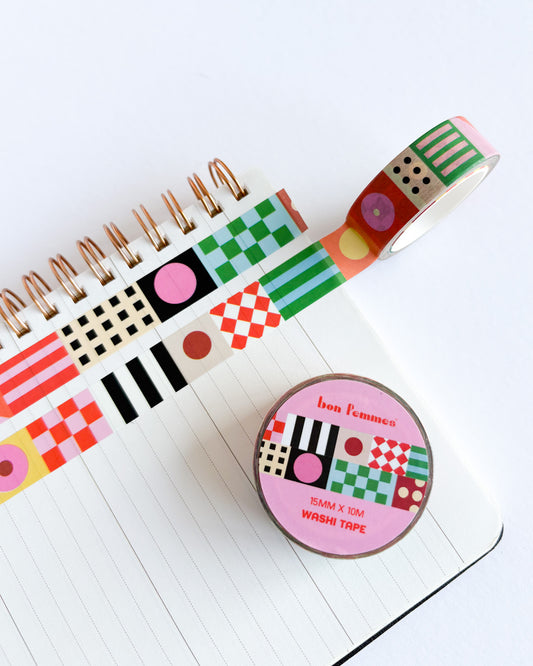 A roll of patterned washi tape and a round tin of Bon Femmes washi tape placed on an open lined notebook. The tape features colorful geometric patterns including stripes, checkers, polka dots, and grids in green, pink, red, black, and white.