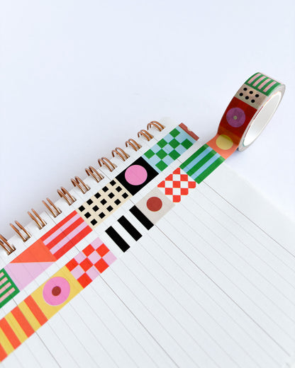 A roll of decorative washi tape with a colorful patchwork pattern of stripes, checkers, dots, and geometric shapes, shown partially unrolled over a white lined spiral notebook on a clean white background.