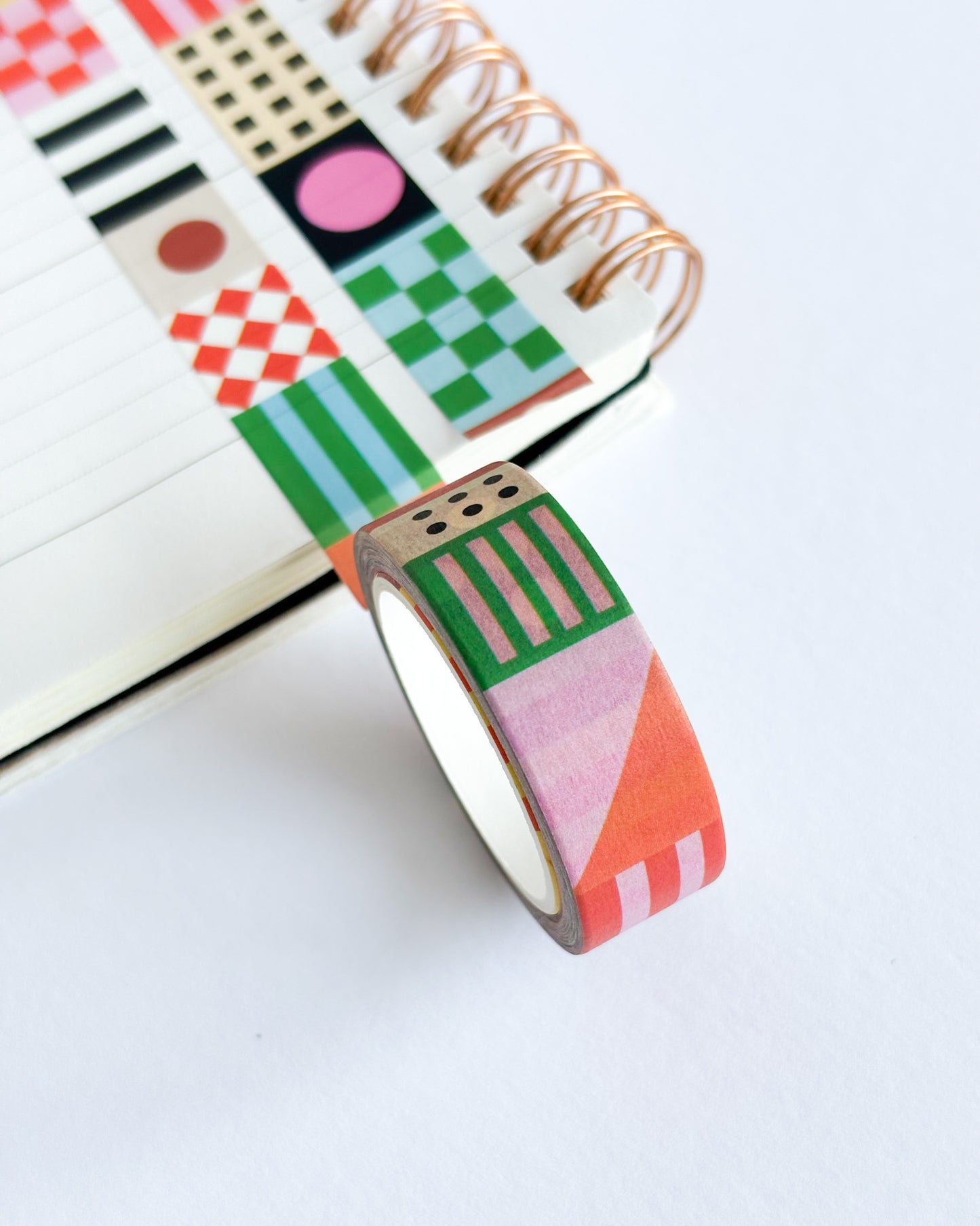 A roll of geometric patterned washi tape in pink, green, orange, and white, shown next to a spiral notebook on a clean white background.