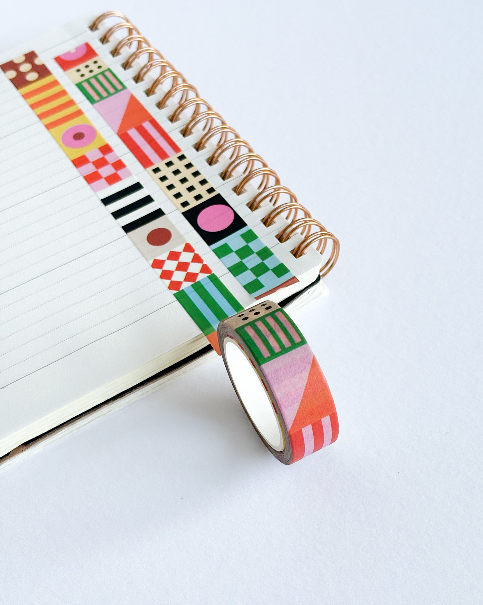 A roll of washi tape with a colorful geometric pattern is draped over an open spiral notebook on a clean white surface.