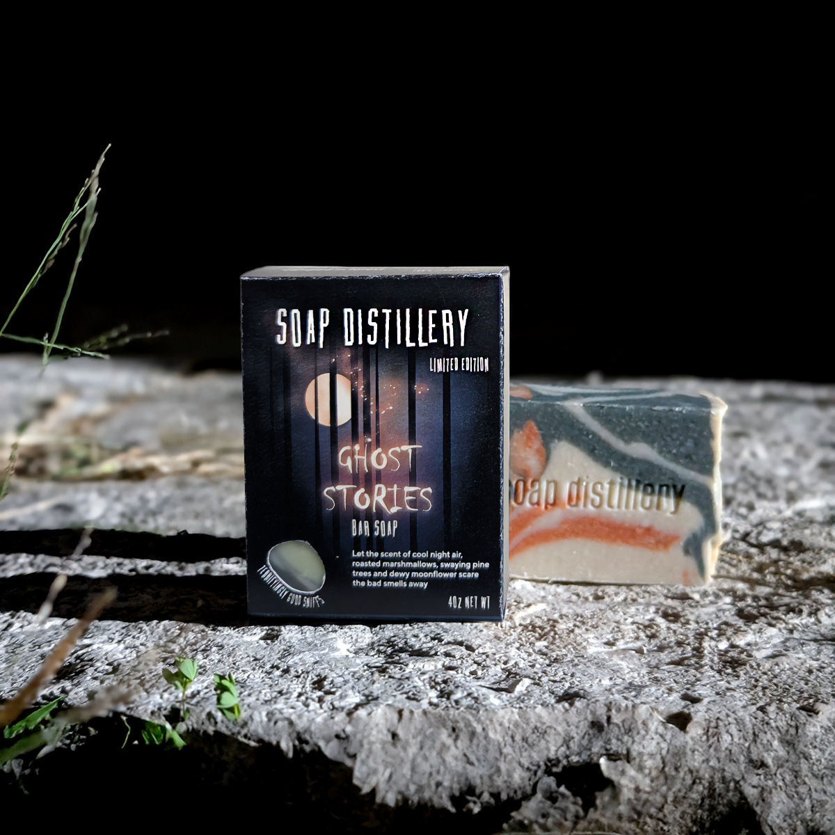 A boxed bar of soap stands in front of a wrapped soap bar on a rough stone surface against a black background. The front box reads 'SOAP DISTILLERY' and 'GHOST STORIES BAR SOAP'; the soap itself shows a marbled green, white, and orange pattern.