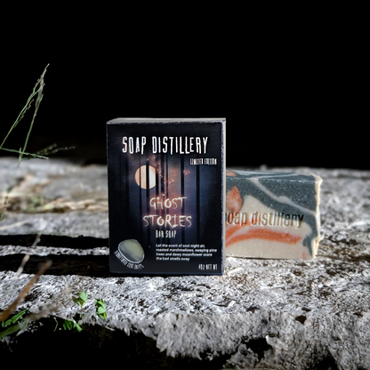 A boxed bar of soap stands in front of a wrapped soap bar on a rough stone surface against a black background. The front box reads 'SOAP DISTILLERY' and 'GHOST STORIES BAR SOAP'; the soap itself shows a marbled green, white, and orange pattern.