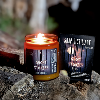 A lit amber glass jar candle labeled 'Ghost Stories' by Soap Distillery sits on a weathered log beside its matching product box. The scene is dark and moody with a blurred outdoor background.