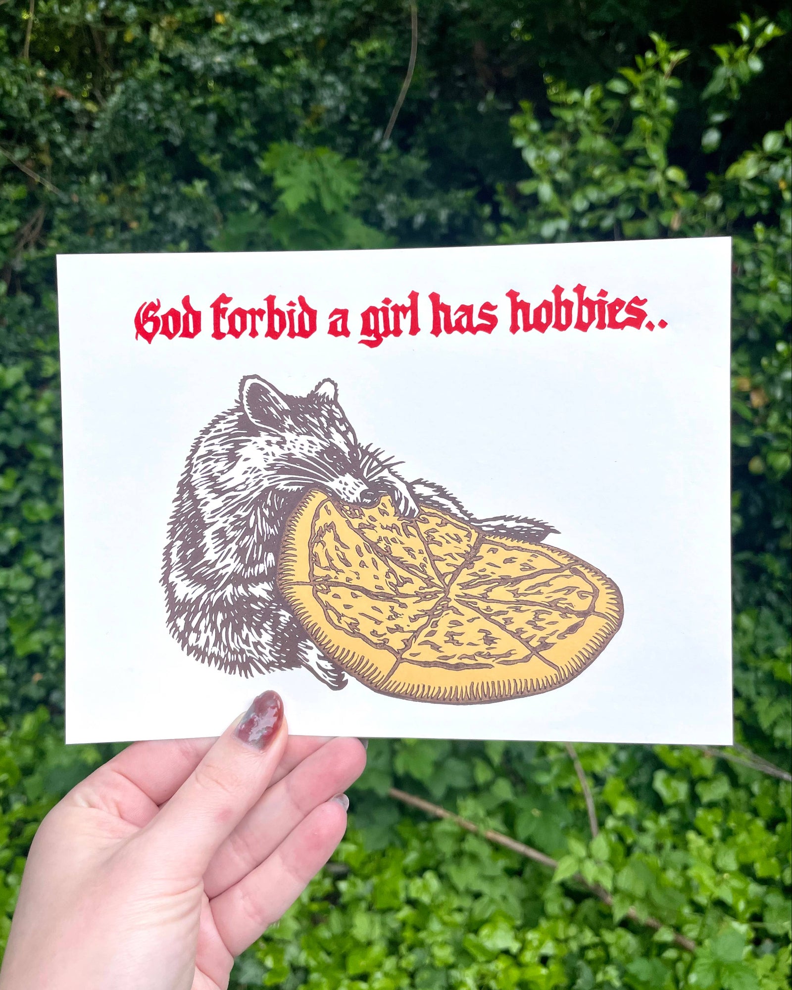 Card with a raccoon eating pizza and text 'God forbid a girl has hobbies' held against a green leafy background.
