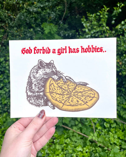 Card with a raccoon eating pizza and text 'God forbid a girl has hobbies' held against a green leafy background.