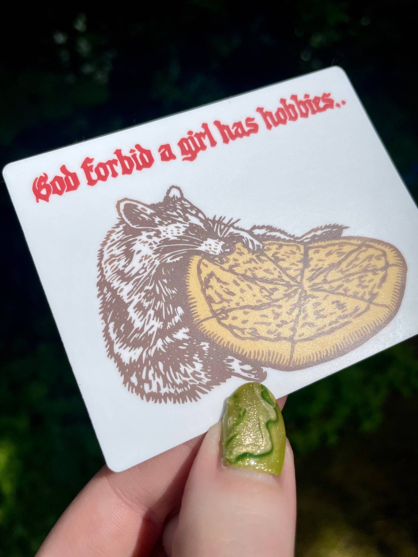 Card with a raccoon and pizza illustration and text held by a hand with green nail polish.