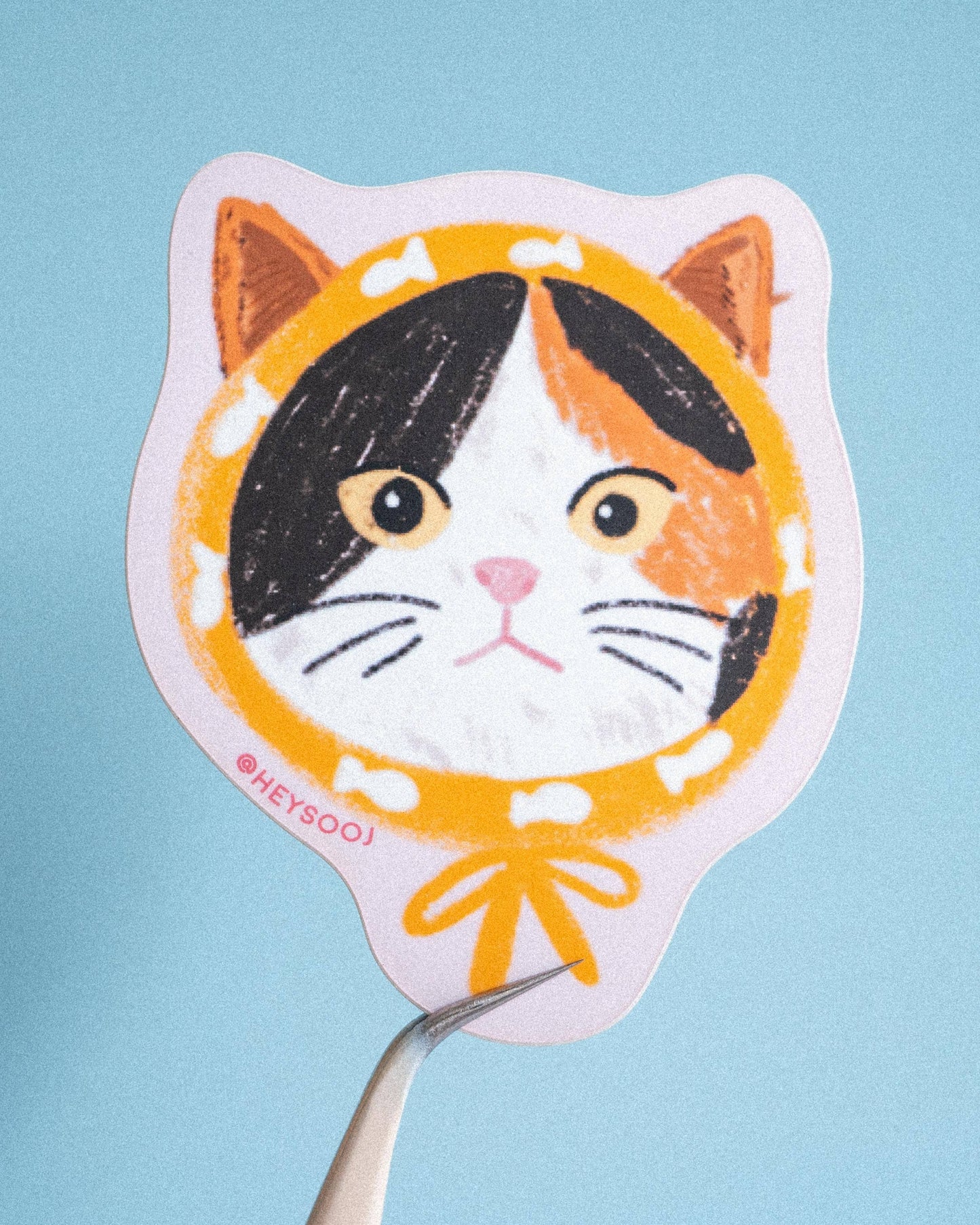 A hand-drawn style sticker or decal of a calico cat face with a yellow collar and bow, held by tweezers against a light blue background.