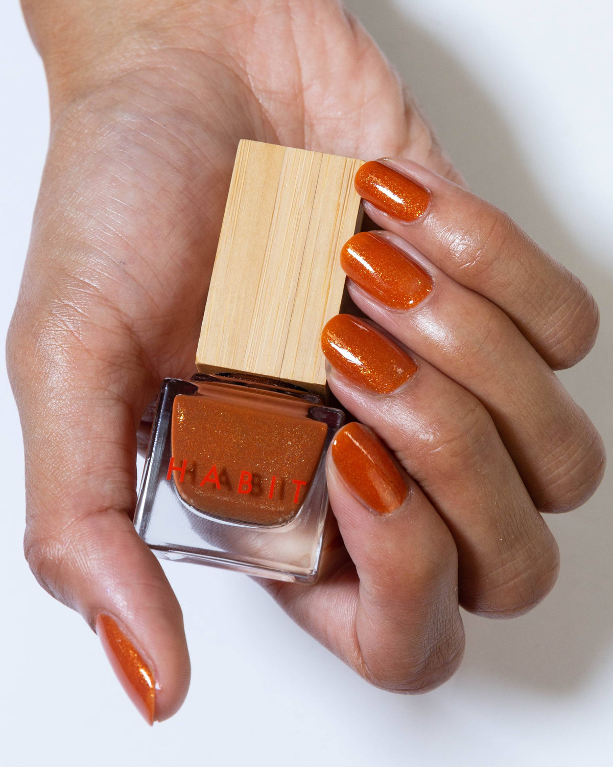 Hand holding a bottle of orange nail polish with a wooden cap on a white , Golden Hour Bronze Shimmer Nail Polishbackground