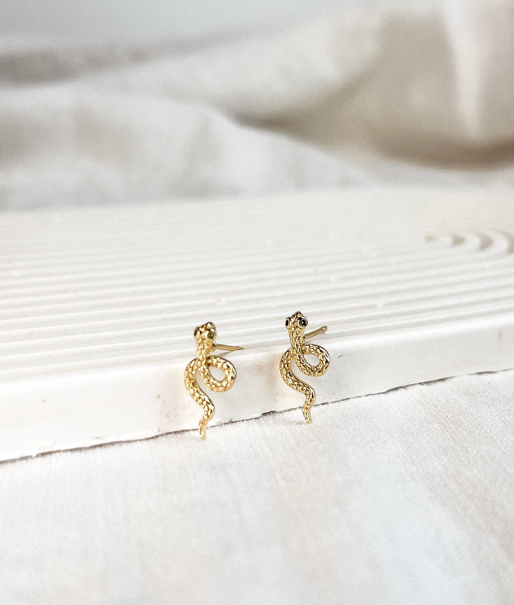 A pair of gold snake-shaped stud earrings resting on a white textured surface with a soft, draped fabric background.