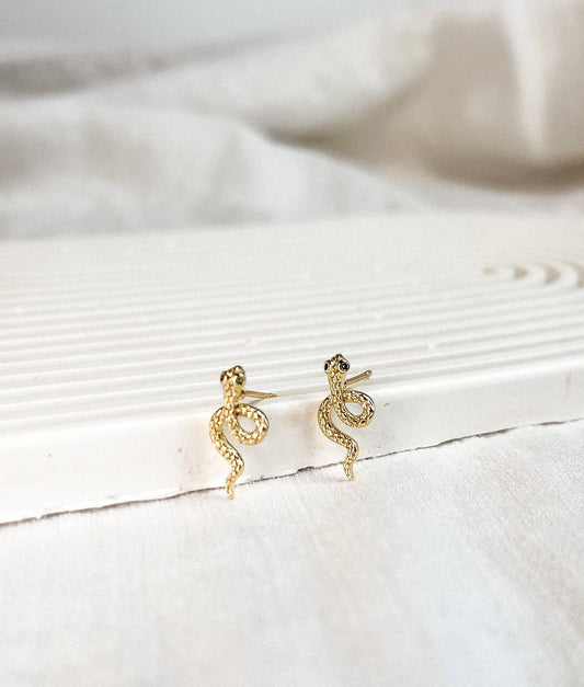 A pair of gold snake-shaped stud earrings resting on a white textured surface with a soft, draped fabric background.