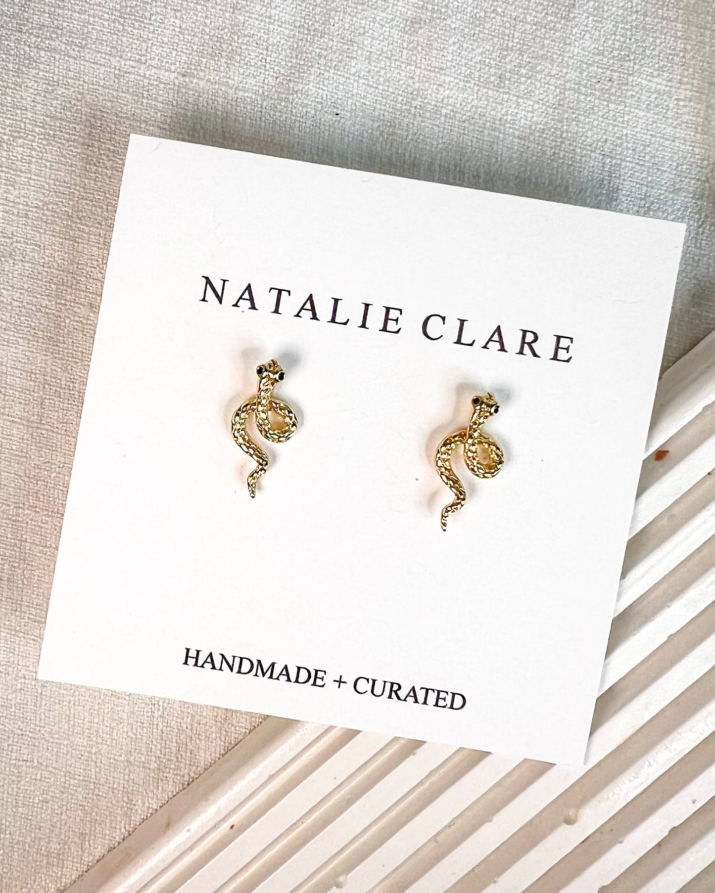 A pair of small gold-toned snake stud earrings displayed on a white Natalie Clare card, placed on a textured neutral fabric surface with a striped prop underneath.