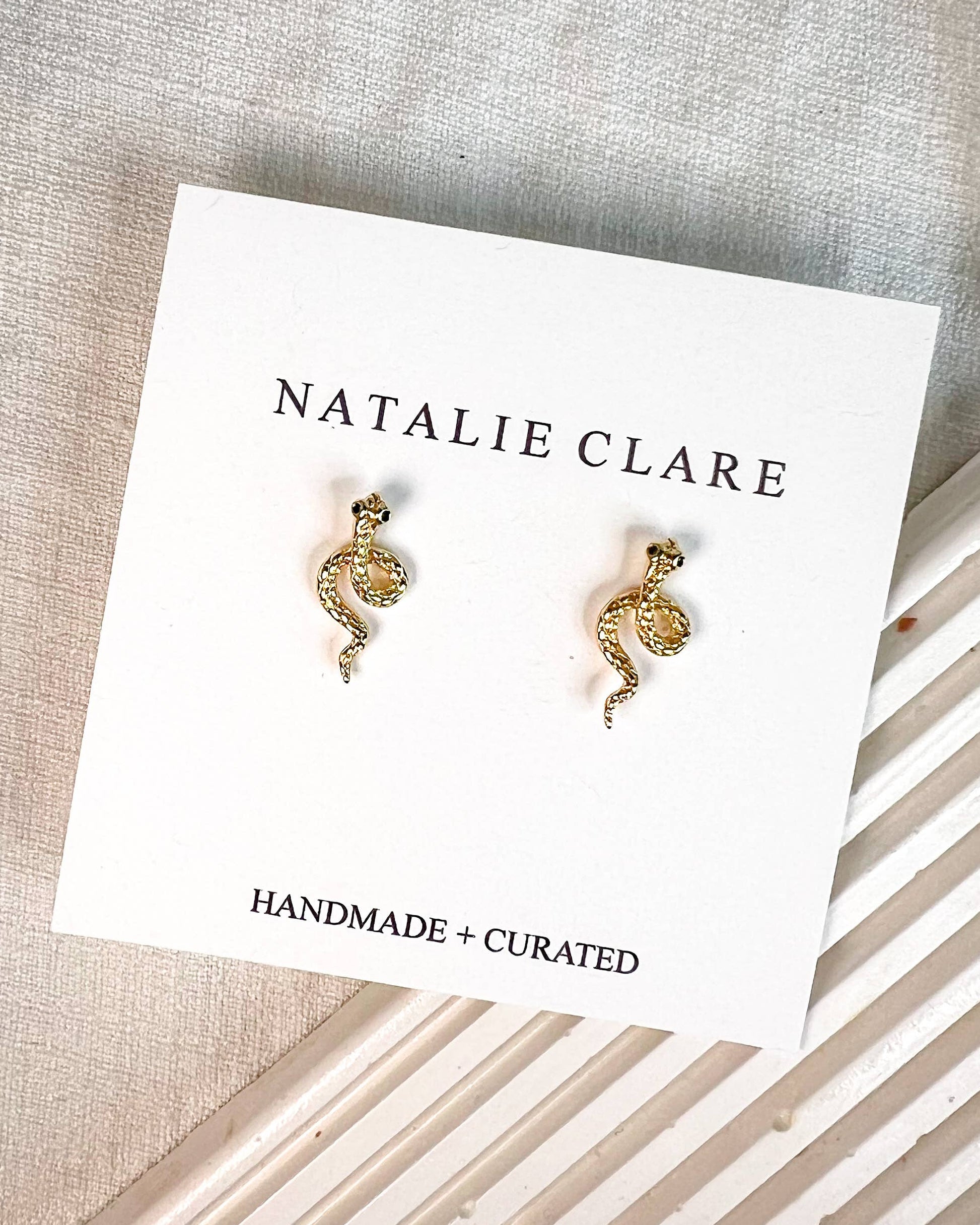 A pair of small gold-toned snake stud earrings displayed on a white Natalie Clare card, placed on a textured neutral fabric surface with a striped prop underneath.