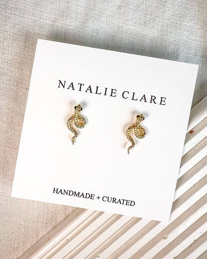 A pair of small gold-toned snake stud earrings displayed on a white Natalie Clare card, placed on a textured neutral fabric surface with a striped prop underneath.