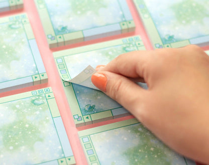A close-up of a hand with orange nail polish peeling back a layer of a printed sticky note from the pad. The printed sheet features a whimsical, retro-style pixel art scene with a green landscape, a small flying saucer, and a light blue sky, framed by pink borders.