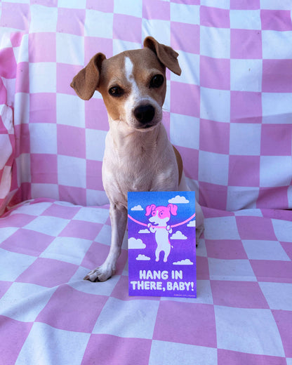 A small brown and white dog sits on a pink and white checkered blanket, holding a purple card that reads 'HANG IN THERE, BABY!' with a cartoon pig.
