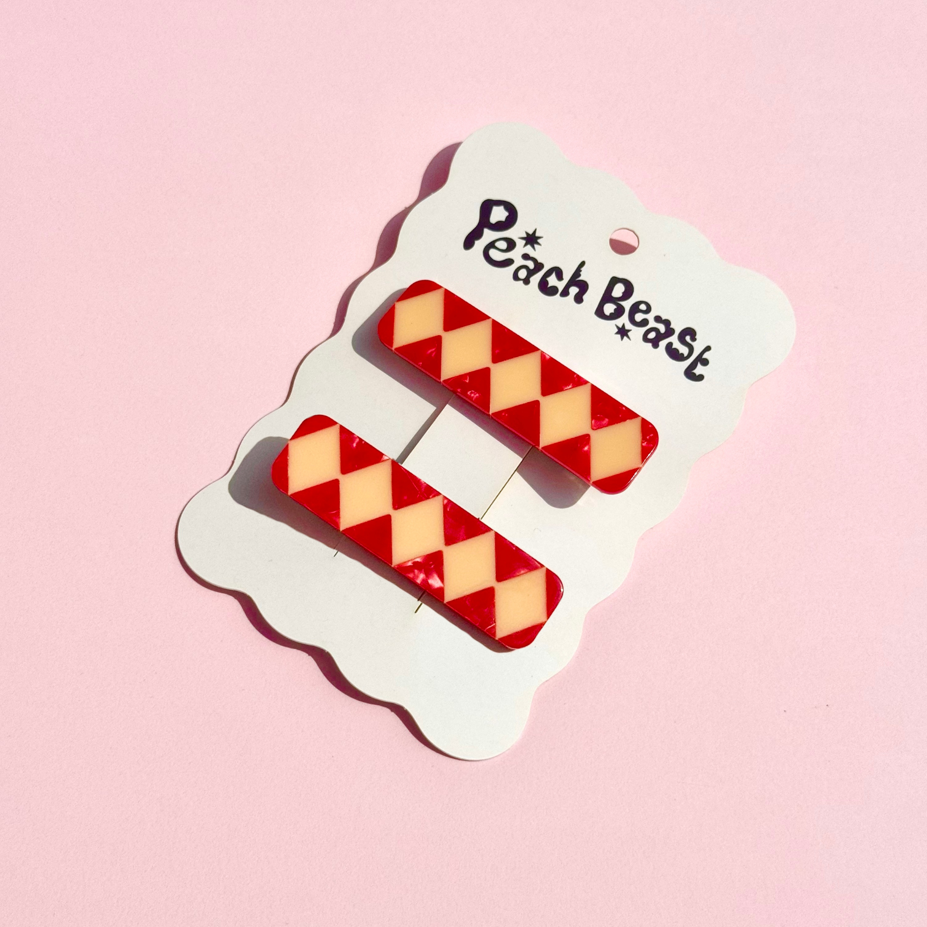 Two red and cream harlequin-patterned hair clips attached to a white cloud-shaped card printed with the brand name 'Peach Beast', set against a solid pink background.