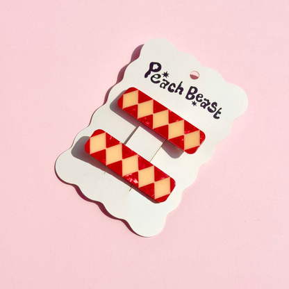 Two red and cream harlequin-patterned hair clips attached to a white cloud-shaped card printed with the brand name 'Peach Beast', set against a solid pink background.