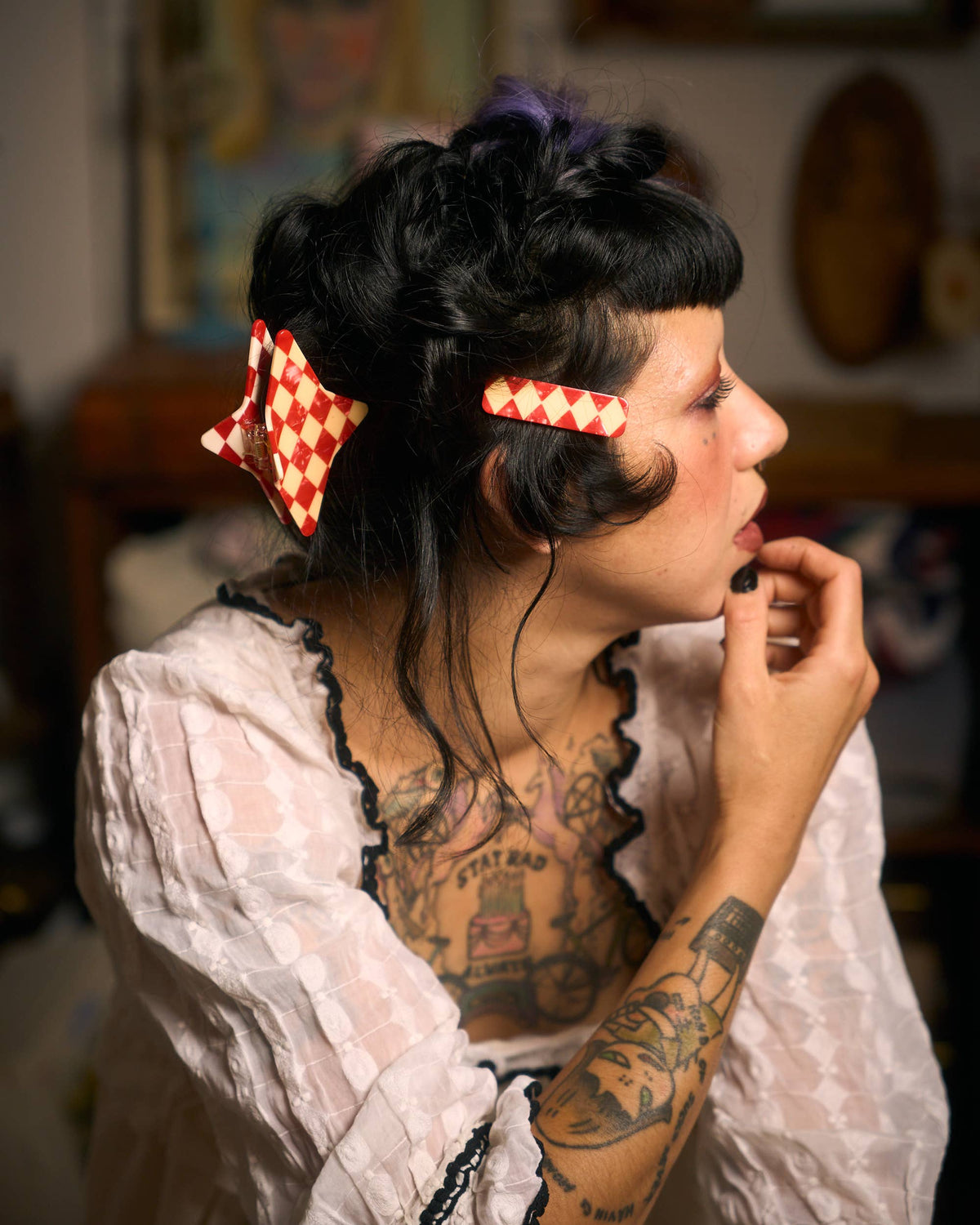 A woman in profile wearing a white lace blouse with black ruffled trim, her dark hair styled in an updo with two red-and-white checkered hair clips. She has visible chest and arm tattoos and is posed indoors against a softly blurred background.