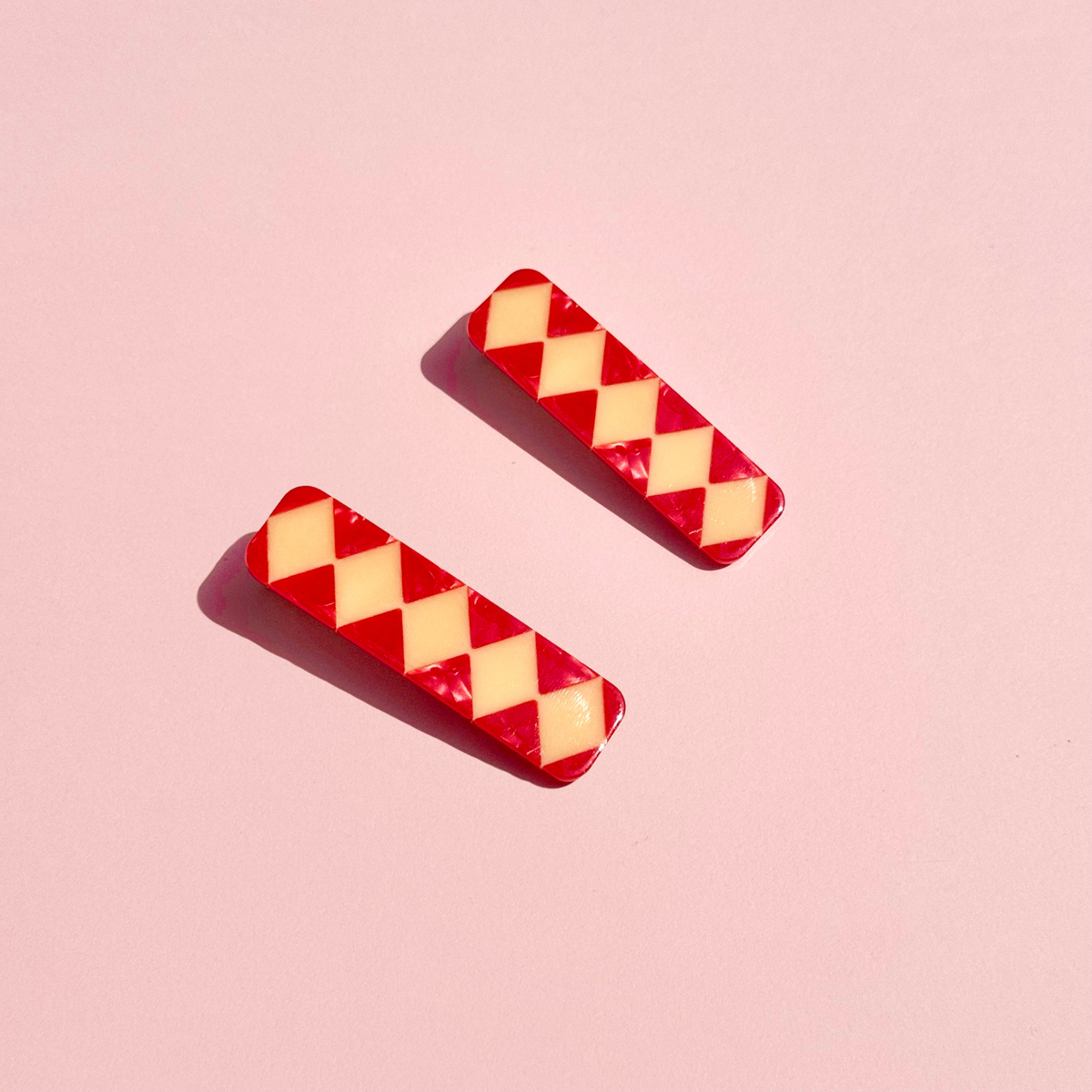 Two rectangular barrettes with a red and pale yellow harlequin diamond pattern, displayed on a solid pink background.