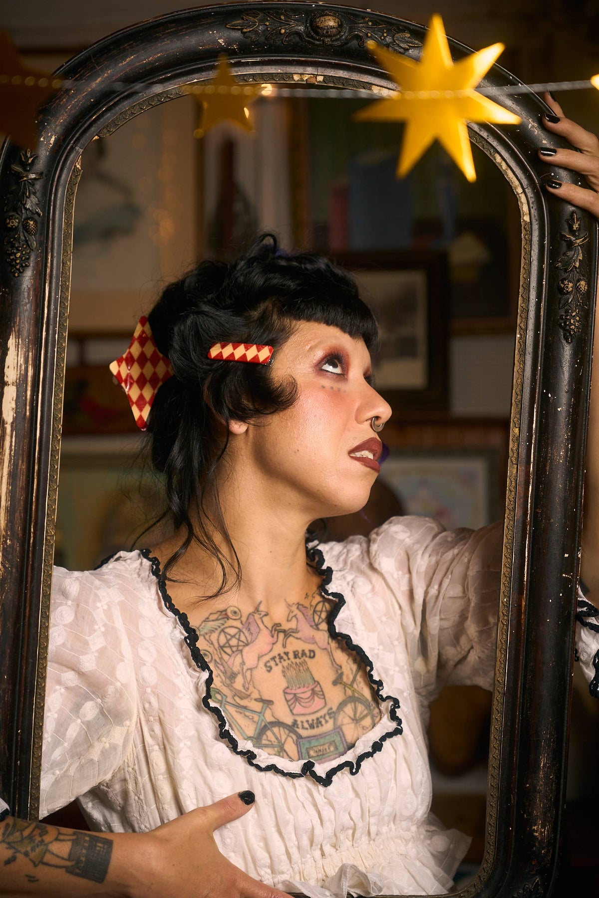 A woman with dark hair and visible chest tattoos looks into an ornate dark mirror, wearing a white ruffled blouse with black trim and red-and-white check accents in her hair.