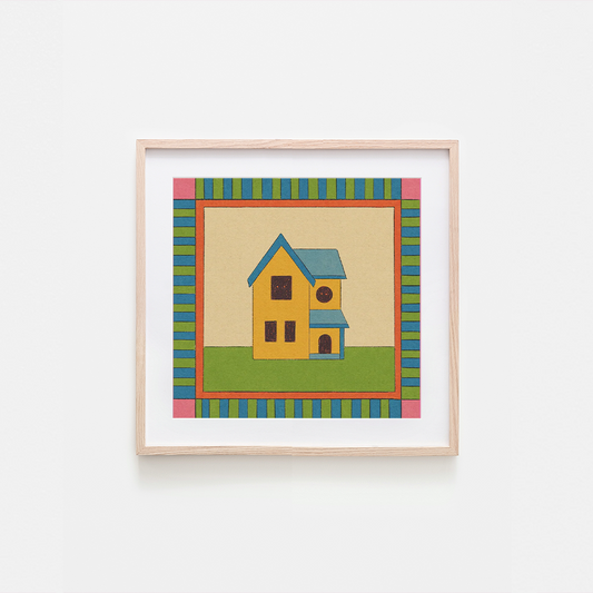 A framed art print featuring a whimsical yellow house with a blue roof, set against a beige background with a colorful striped border, hung on a plain white wall.