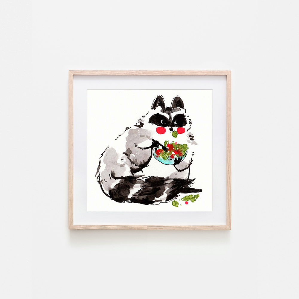A framed art print featuring a whimsical watercolor illustration of a raccoon with black and white fur, red cheeks, and a bowl of salad, displayed on a light gray wall.