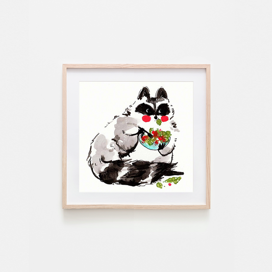 A framed art print featuring a whimsical watercolor illustration of a raccoon with black and white fur, red cheeks, and a bowl of salad, displayed on a light gray wall.