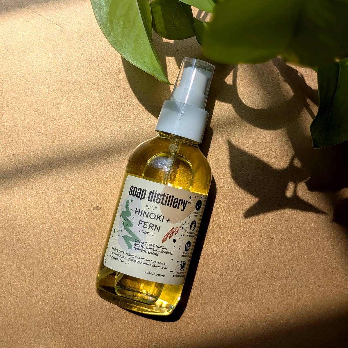 Bottle of Soap Distillery Hinoki Fern oil on a beige surface with green leaves.