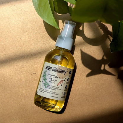 Bottle of Soap Distillery Hinoki Fern oil on a beige surface with green leaves.