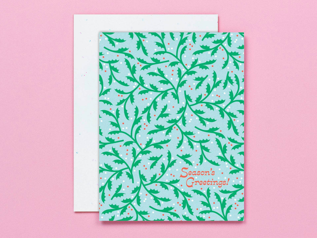 A square greeting card with a light blue background and a green holly pattern, with the words 'Season's Greetings!' in orange script. A white envelope is partially visible behind it on a solid pink background.