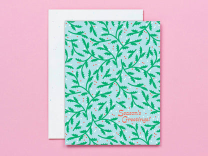 A square greeting card with a light blue background and a green holly pattern, with the words 'Season's Greetings!' in orange script. A white envelope is partially visible behind it on a solid pink background.