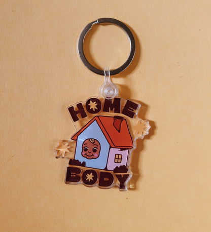 A custom acrylic keychain featuring a cartoon house with a baby's face in the window, surrounded by the words 'HOME BODY' in brown letters, on a warm beige background.