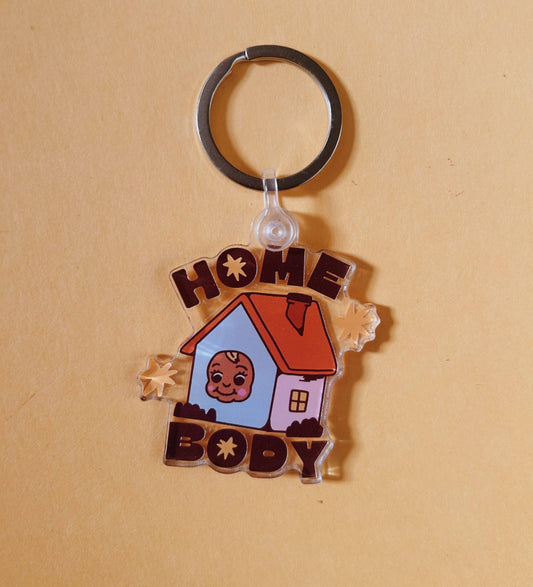 A custom acrylic keychain featuring a cartoon house with a baby's face in the window, surrounded by the words 'HOME BODY' in brown letters, on a warm beige background.