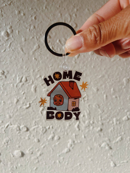 A hand holding a clear acrylic keychain with a black split ring. The keychain features a cartoon house with a smiling face, the words 'HOME BODY' in dark brown, and small orange starburst accents, set against a textured off-white wall.