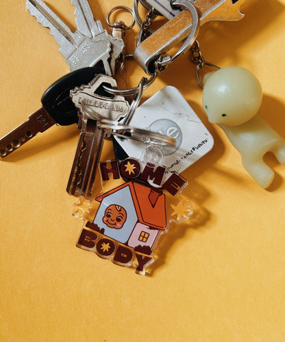 A close-up of a keychain set on a warm yellow background. The set includes silver keys, a small pale green figure, and a colorful acrylic charm shaped like a house with the words “HOME BODY” and a cartoon face.