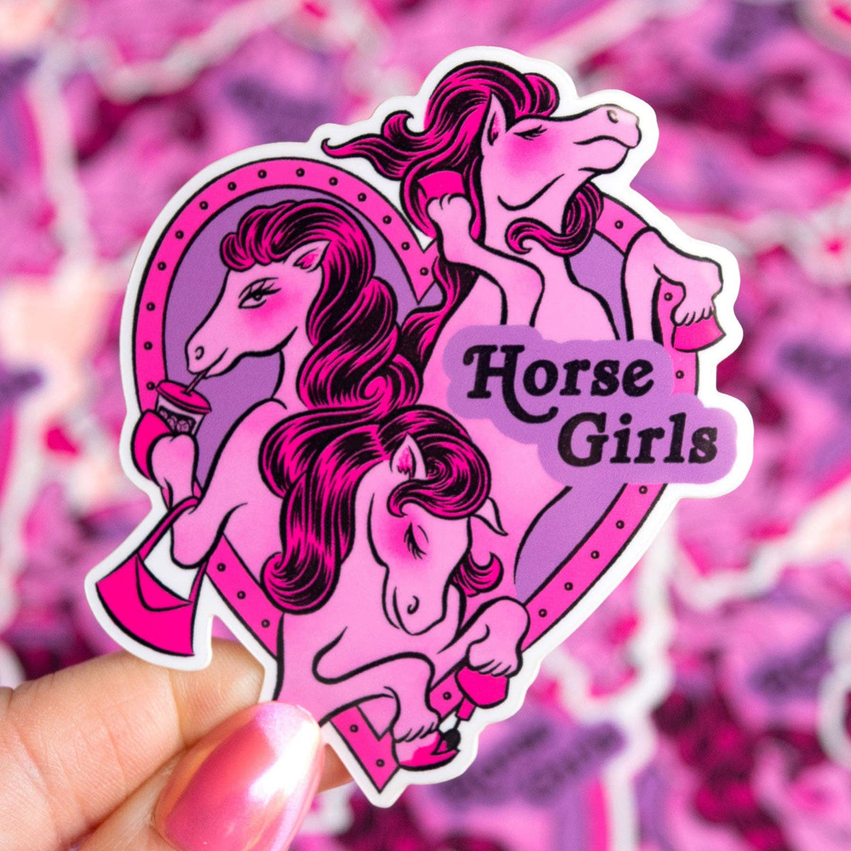 A die-cut sticker featuring three stylized pink and purple horse girls, held by a hand with pink nail polish against a blurred pink patterned background. The sticker includes the text 'Horse Girls'.